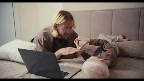 A-happy-blond-man-in-glasses-with-a-beard-plays-with-his-little-albino-son-with-white-hair-color-in-rock-paper-scissors-and-then-turns-on-a-cartoon-on-a-gray-laptop-while-spending-time-together-in-the-room