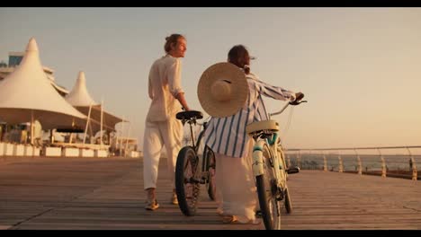 Rear-view-of-a-guy-and-a-girl-in-light-beachwear-Walking-with-their-bicycles-along-the-beach-covered-with-boards-along-the-sea-at-Sunrise-in-summer