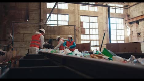 Side-view-of-waste-recycling-plant-workers-in-special-uniforms-laying-out-and-sorting-plastic-bottles-based-on-their-color-while-standing-near-a-moving-conveyor-belt-at-a-big-old-plant