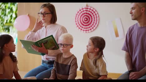 A-blonde-woman-with-a-boxy-hairstyle-and-a-pink-shirt-reads-a-green-book-to-preschool-children-in-a-special-club-for-preparing-children-for-school