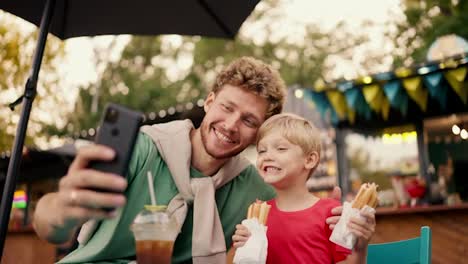 Glücklicher-Vater-Mit-Lockigem-Haar-Und-Grünem-T-Shirt-Macht-Ein-Selfie-Mit-Seinem-Kleinen-Sohn-Mit-Blauen-Augen-In-Einem-Roten-T-Shirt,-Der-Zwei-Hot-Dogs-In-Den-Händen-Hält,-Während-Er-Auf-Einem-Straßentisch-In-Einem-Straßencafé-Im-Park-Sitzt