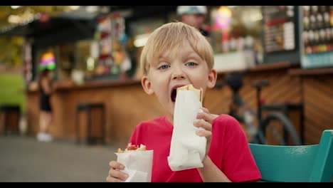 Porträt-Eines-Glücklichen-Kleinen-Blonden-Jungen-Mit-Blauen-Haaren-In-Einem-Roten-T-Shirt,-Der-Einen-Hot-Dog-Isst-Und-Hot-Dogs-In-Den-Händen-Hält,-Während-Er-An-Einem-Straßentisch-In-Einem-Café-Im-Park-Sitzt