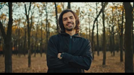 Fröhlicher-Und-Glücklich-Lächelnder-Mann-Mit-Mittellangem-Haar-Und-Brünettem-Bart-In-Schwarzer-Sportuniform-Posiert-Im-Herbstlichen-Wald