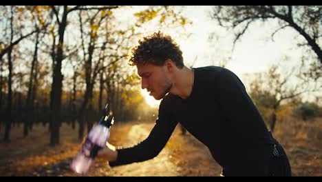 A-happy-and-tired-man-with-curly-hair-in-a-black-sports-uniform-pours-water-from-a-sports-bottle-on-his-face-and-refreshes-himself-after-a-hard-jog-in-an-autumn-park-in-the-morning