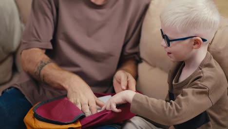 Close-up-shot-of-a-blond-man-with-a-beard-and-glasses-helping-his-little-albino-son-with-white-hair-in-blue-glasses-pack-his-backpack-before-going-to-school-in-the-morning