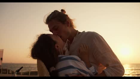 Feliz-Pareja,-Un-Chico-Con-Ropa-Clara-Y-Una-Chica-Con-Una-Camisa-Blanca-Y-Azul-Besándose-Al-Amanecer-Cerca-Del-Mar.-Feliz-Pareja-En-Una-Cita-En-La-Playa-Cerca-Del-Mar-Al-Amanecer-En-Verano