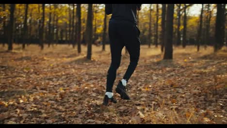 Un-Hombre-Con-Un-Uniforme-Deportivo-Negro,-Cabello-Rizado-Y-Zapatillas-Deportivas-Corre-Por-Un-Bosque-Otoñal-Con-Hojas-Caídas-En-Un-Día-Soleado.-Vista-Lateral-De-Un-Joven-Atleta-Feliz-Corriendo-Por-El-Bosque-Otoñal-En-Una-Soleada-Mañana-De-Otoño.