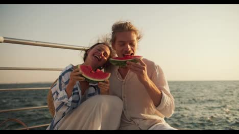 A-happy-couple-in-love:-a-blond-guy-with-stubble-in-light-clothes-and-a-brown-haired-girl-sitting-on-a-pier-near-the-sea-and-eating-watermelon-on-a-picnic.-A-guy-and-a-girl-eat-juicy-watermelon-slices-on-a-picnic-near-the-sea