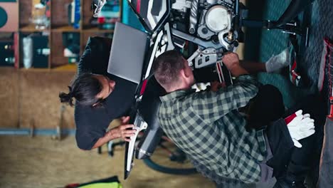 Vertical-video:-2-mechanics-repairing-a-bike.-A-brunette-man-in-a-gray-T-shirt-looks-at-the-screen-of-a-gray-laptop-and-a-man-in-a-checkered-shirt-repairs-a-motorcycle-in-a-workshop-garage