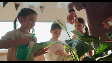 A-preschool-girl-uses-a-special-green-sprayer-to-spray-a-flower-with-water-and-water-it-under-the-attention-of-her-teacher.-A-man-in-a-purple-T-shirt-during-a-break-at-a-club-to-prepare-children-for-school