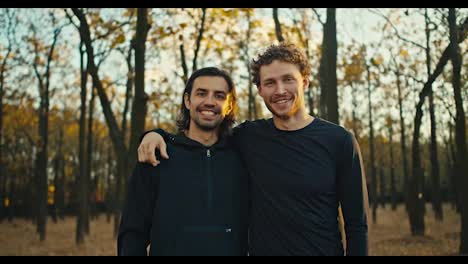 Portrait-of-two-happy-guys-with-black-hair-and-beard-in-black-sportswear-who-are-on-a-morning-jog-in-the-autumn-forest-in-the-morning.-Two-guys-are-hugging-and-posing-in-the-morning-In-the-forest