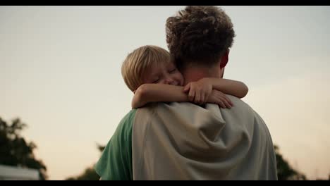 A-little-blond-boy-lovingly-hugs-the-neck-of-his-dad-with-curly-hair-in-a-green-T-shirt.-Little-boy-loves-his-dad-and-hugs-him