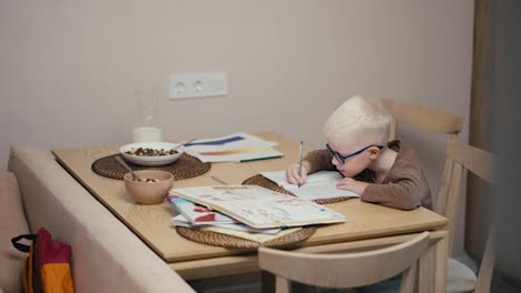 Pequeño-Niño-Albino-Con-Cabello-Blanco-Y-Gafas-Azules-Haciendo-Los-Deberes-Y-Escribiendo-En-Sus-Cuadernos-Mientras-Se-Prepara-Para-El-Día-Siguiente-En-La-Escuela-En-Una-Cocina-Moderna-En-Un-Apartamento-Durante-El-Día.