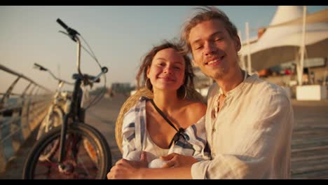 Portrait-of-a-happy-couple:-a-blond-guy-with-stubble-in-a-light-shirt-and-a-girl-with-earrings-relax-on-the-beach,-which-is-covered-with-wooden-boards,-against-of-white-cafe-umbrellas.-Happy-vacation-at-sea-during-a-slight-wind,-a-guy-and-a-girl-rode-a-bicycle-to-the-beach