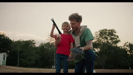 Portrait-of-a-Happy-blond-boy-in-a-red-T-shirt-who-holds-a-baseball-bat-and-stands-with-his-dad-in-a-Green-T-shirt-with-curly-hair-and-stubble-and-posing-in-the-park