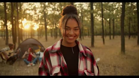 Portrait-of-a-happy-girl-with-black-skin-in-a-checkered-shirt-who-looks-at-the-camera-and-poses-against-the-background-of-other-participants-in-a-hike-and-tent-in-a-Sunny-summer-green-forest