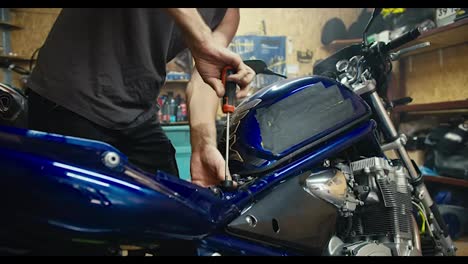 Close-up-shot:-a-man-in-a-gray-T-shirt,-a-mechanic,-in-a-gray-T-shirt,-repairs-a-blue-motorcycle-with-a-screwdriver-in-his-garage-workshop