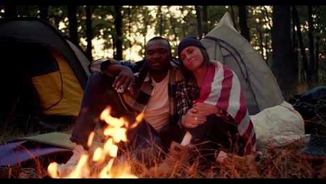 A-man-with-black-skin-and-a-girl-with-a-bob-in-a-black-hat-sit-near-a-fire-during-a-camping-stop-and-hug-under-the-US-flag-against-the-backdrop-of-a-beautiful-forest-and-tents