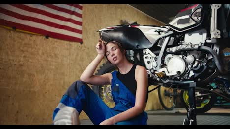 Una-Chica-Mecánica-De-Automóviles-Cansada,-Con-Una-Camiseta-Negra-Y-Un-Mono-Azul,-Se-Apoya-En-Una-Motocicleta-Que-Levantó-Y-Descansa-En-Su-Taller-Con-Una-Bandera-Estadounidense-En-La-Pared.