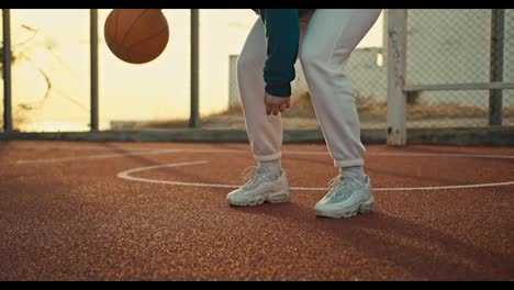 Primer-Plano-De-Una-Niña-Con-Zapatillas-Blancas-Y-Uniforme-Deportivo-Golpea-Una-Pelota-Naranja-Desde-El-Suelo-En-La-Cancha-De-Baloncesto-De-La-Calle-Roja-Por-La-Mañana.