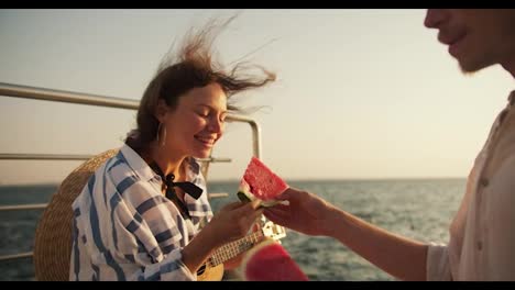 The-guy-invites-the-girl-to-eat-a-piece-of-watermelon-at-a-picnic-on-the-pier-near-the-sea.-The-girl-bites-the-watermelon-and-rejoices.-Happy-couple-on-a-date-near-the-sea-on-the-pier