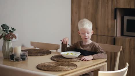 Kleiner-Albinojunge-Mit-Weißem-Haar-Isst-Haferbrei-Mit-Weintrauben,-Während-Er-Auf-Einem-Holzstuhl-In-Einer-Modernen-Küche-Sitzt-Und-Frühstückt.-Kleiner-Albinojunge-Mit-Weißem-Haar-Frühstückt-In-Der-Küche