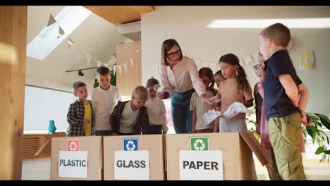 Eine-Lehrerin-Mit-Bob-Frisur,-Brille-Und-Weißem-Hemd-Bringt-Kindern-In-Einem-Vorschulclub-Bei,-Wie-Man-Müll-Richtig-Sortiert,-Um-Sie-Auf-Die-Schule-Vorzubereiten.-Ein-Blondes-Mädchen-Zeigt-Den-Kindern,-Wie-Man-Müll-Richtig-Sortiert-Und-In-Welche-Kisten-Sie-Nach-Kategorien-Werfen.