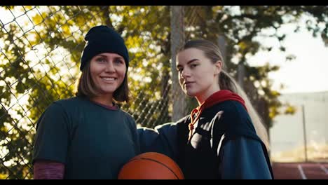 Portrait-of-two-happy-girls-in-sportswear-with-a-basketball-orange-ball-on-a-street-sports-field-in-the-morning-on-a-sunny-summer