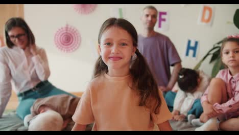 a-happy-girl-with-blue-eyes-in-an-orange-T-shirt-looks-at-the-camera-and-smiles-against-the-background-of-her-first-lesson-with-teachers-in-preparation-for-school.-Preschool-girl-preparing-for-school-with-her-teachers-and-a-group-of-children