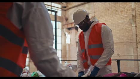 Shooting-close-up-a-man-with-Black-skin-in-a-special-white-protective-uniform-together-with-his-colleague-sorts-and-sorts-garbage-at-a-large-waste-processing-plant-near-a-conveyor-belt