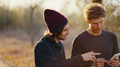 Close-up-shot-of-two-brunette-athlete-friends-in-a-black-sports-uniform-walking-and-talking-while-looking-at-something-on-the-smartphone-screen-during-their-walk-along-an-earthly-path-in-a-sunny-autumn-forest-in-the-morning