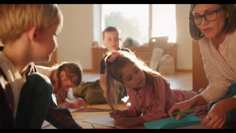 A-woman-teacher-in-a-special-club-for-preparing-children-for-school,-telling-her-children-how-to-draw-correctly-and-conducting-a-drawing-lesson-on-the-floor-children-drawing-on-multi-colored-paper-using-pencils-in-a-cozy-sunny-room-with-large-windows