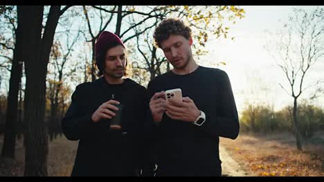 A-confident-and-happy-man-with-curly-hair-in-a-black-sports-uniform-with-a-white-phone-in-his-hands-walks-and-communicates-with-his-friend-a-man-with-a-red-during-their-walk-in-the-autumn-forest-in-the-evening