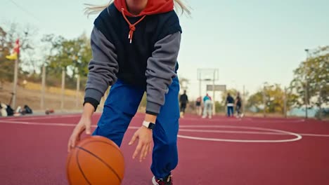 Primer-Plano-De-Una-Chica-Rubia-Con-Uniforme-Deportivo-Que-Corre-Y-Maniobra-Con-Una-Espada-Naranja-Y-Lanza-La-Pelota-Al-Aro-Durante-Su-Juego-De-Baloncesto-En-La-Cancha-De-La-Calle.