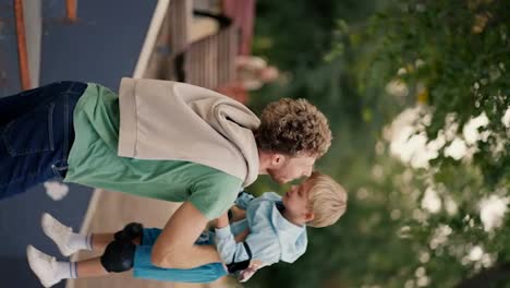 Vertical-video-of-a-dad-in-a-Green-T-shirt-throwing-his-little-blond-son-in-blue-clothes-on-a-skate-park-in-the-summer-in-the-park