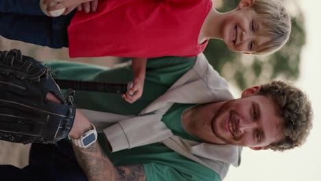 Vertical-video:-A-dad-with-curly-hair-with-stubble-in-a-Green-T-shirt-with-a-baseball-glove-on-his-hand-and-near-his-little-blond-son-in-a-red-T-shirt-with-a-baseball-bat-pose-together-on-the-playground-in-the-park