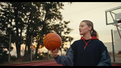 Ein-Glückliches-Blondes-Mädchen-In-Einem-Schwarzen-Kapuzenpullover-Versucht,-Einen-Orangefarbenen-Basketball-Auf-Ihrem-Zeigefinger-Zu-Drehen,-Während-Sie-Am-Frühen-Morgen-Auf-Einem-Roten-Basketballplatz-Basketball-Trainiert