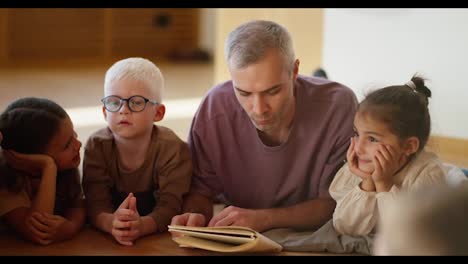 A-male-teacher-with-gray-hair-in-a-purple-T-shirt-reads-a-book-to-preschool-children-who-lie-on-the-floor-on-special-pillows-in-a-club-for-preparing-children-for-school-in-a-cozy-room