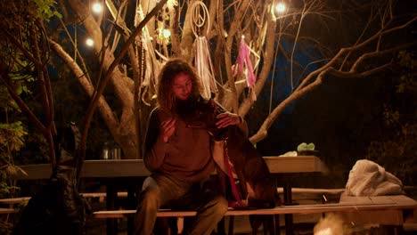 A-curly-haired-boy-feeds-a-black-dog-on-a-bench-near-a-campfire-against-a-forest-background.-Rest-in-the-country-house