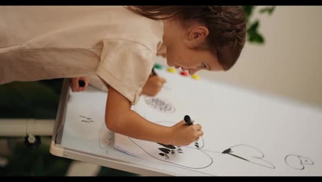 Vertical-video-of-a-girl-in-a-white-T-shirt-drawing-with-a-black-marker-on-a-large-sheet-of-paper-that-is-installed-on-a-magnetic-board-in-the-room-of-the-club-for-preparing-preschoolers-for-school