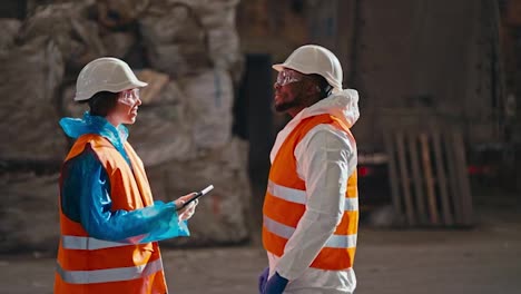 A-brunette-girl-in-a-blue-uniform-and-an-orange-vest-communicates-with-a-man-with-Black-skin-in-a-white-uniform-while-they-are-at-a-large-waste-processing-and-sorting-plant