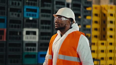 Un-Hombre-De-Piel-Negra-Con-Un-Uniforme-De-Protección-Blanco-Y-Un-Chaleco-Naranja-Con-Gafas-De-Seguridad-Transparentes-Camina-Junto-A-Cajas-Amarillas-Y-Negras-En-Una-Planta-De-Procesamiento-Y-Clasificación-De-Residuos.