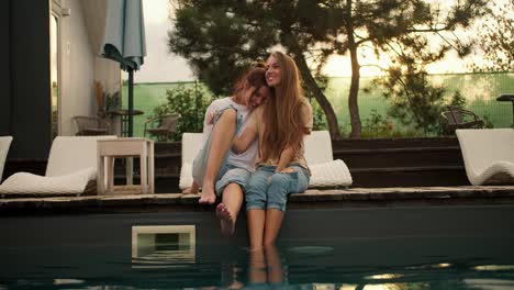 Two-girlfriends-are-sitting-near-the-sunbeds-by-the-pool,-hugging-and-looking-into-the-distance.-Rest-in-the-country-house