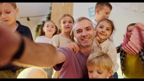 Happy-male-teacher-with-gray-hair-in-a-purple-t-shirt-takes-a-selfie-with-his-students-in-a-club-for-preparing-children-for-school.-The-preschool-children-posing-with-their-teacher