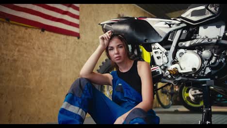 Portrait-of-a-tired-brunette-girl-auto-mechanic-in-a-black-top-and-blue-overalls-who-folds-her-hands-on-her-knees-leaning-on-which-she-has-jacked-up-and-is-repairing-in-a-workshop-with-a-USA-flag-on-the-wall