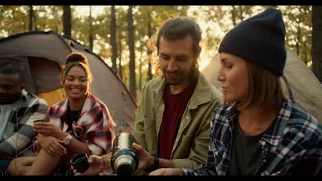 A-happy-group-of-four-hiking-participants-are-developing-and-drinking-tea-from-a-thermos-against-the-backdrop-of-tents-in-a-sunny-green-summer-forest.-An-international-group-of-hikers-in-a-sunny-summer-forest