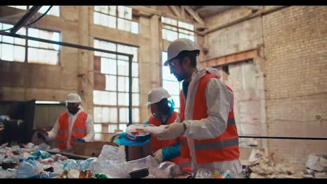 Eine-Gruppe-Von-Drei-Arbeitern-In-Weißen-Uniformen-Und-Orangefarbenen-Westen-Recycelt-Müll-Und-Plastik-Auf-Einem-Förderband-In-Einer-Abfallrecyclinganlage.-Prozess-Der-Sortierung-Von-Flaschen-Nach-Farbe-In-Einer-Abfallrecyclinganlage