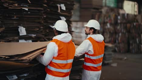 A-man-with-Black-skin-in-a-white-protective-uniform-in-an-orange-vest-together-with-his-colleague-a-man-with-a-beard-lifts-a-large-rack-of-cardboard-at-a-waste-recycling-and-sorting-plant-for-waste-paper-and-plastic