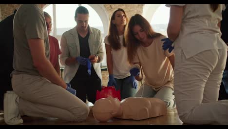 Close-up-shooting:-A-girl-in-a-white-T-shirt-attaches-a-medical-electrical-sensor-to-the-body-of-a-mannequin-under-the-guidance-of-a-professional-doctor,-a-girl-in-a-white-uniform,-while-other-members-of-the-group-listen-to-the-doctor's-explanations.-First-aid-training-against-a-backdrop-of-white-br