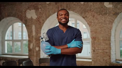 Portrait:-A-professional-Black-male-doctor-in-a-blue-medical-uniform-stands-and-smiles-against-the-background-of-brick-walls-and-large-white-windows